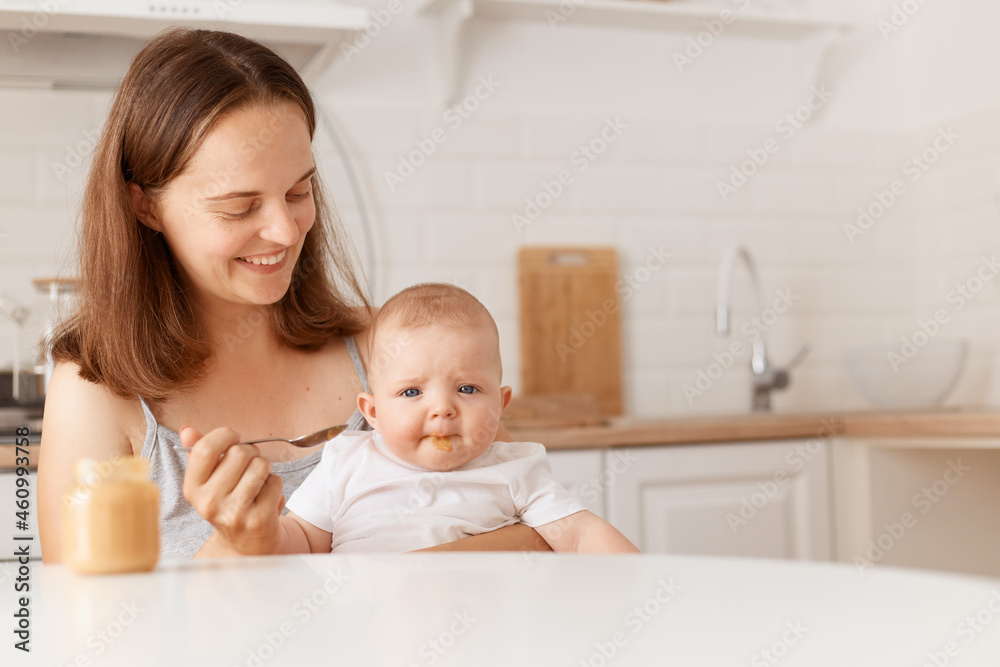 Smiling dark haired female feeds her little daughter with fruit or vegetable puree, sitting in light room with kitchen set on background, first feeding up.