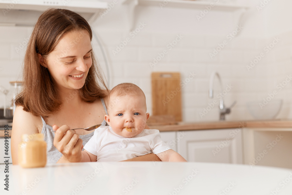 Smiling dark haired female feeds her little daughter with fruit or vegetable puree, sitting in light room with kitchen set on background, first feeding up. Smiling dark haired female feeds her little daughter with fruit or vegetable puree, sitting in light room with kitchen set on background, first feeding up.