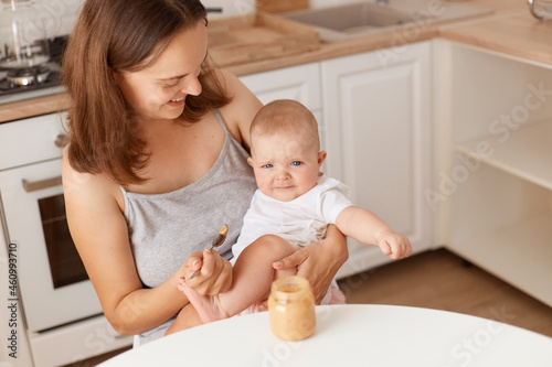 Indoor shot of dark haired female feeds her little daughter with fruit or vegetable puree, mother looking at her cute baby with love, healthy feeding up.