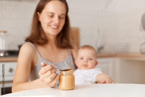 Positive smiling young adult dark haired mother feeding her little daughter with fruit or vegetable puree, holding spoon with healthy food, posing in kitchen.