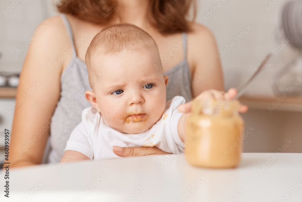 Closeup portrait of cute adorable kid wearing white t shirt, touching jar with food with interest, faceless mother feeding her little infant daughter with vegetable puree. Closeup portrait of cute adorable kid wearing white t shirt, touching jar with food with interest, faceless mother feeding her little infant daughter with vegetable puree.