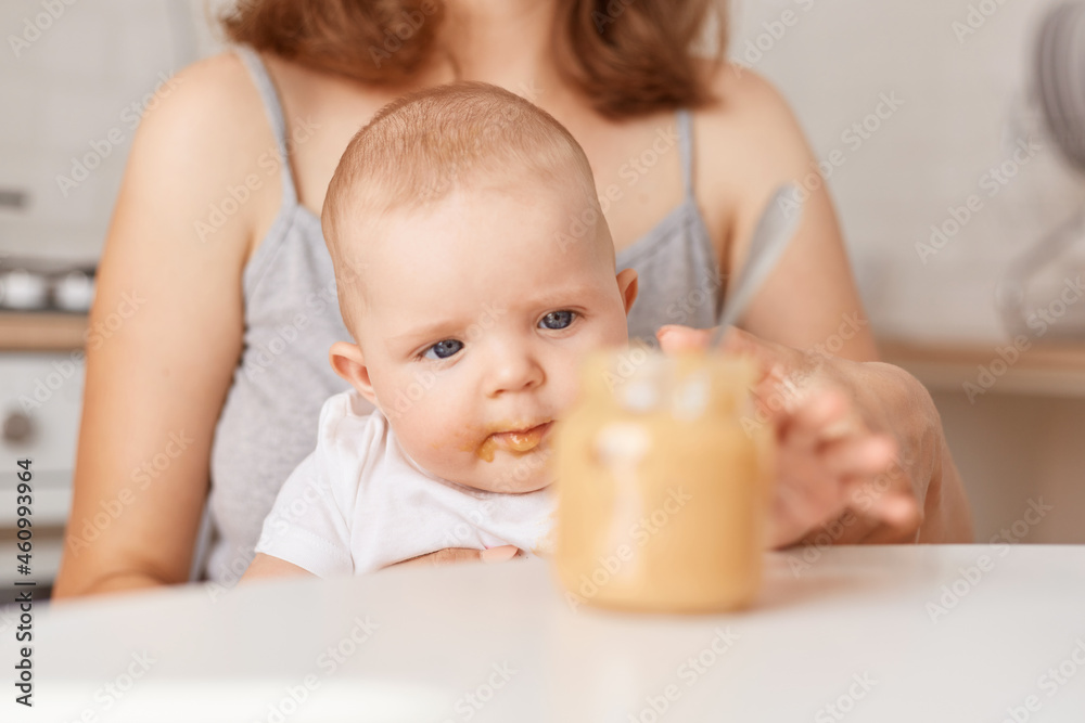 Indoor shot of cute baby in white t shirt sitting with mommy, unknown woman holding feeding infant baby with puree, posing in light kitchen at home, parenthood.