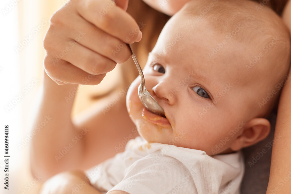 Indoor shot of unknown female holding feeding infant baby, cute charming adorable toddler looking at camera, eating fruit or vegetable puree, mommy taking care of daughter. Indoor shot of unknown female holding feeding infant baby, cute charming adorable toddler looking at camera, eating fruit or vegetable puree, mommy taking care of daughter.