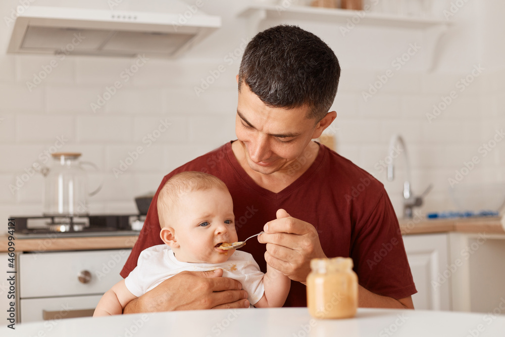 Indoor shot of brunette man feeding little daughter with fruit or vegetable puree while sitting at table in kitchen, people posing at home, single father taking care of baby. Indoor shot of brunette man feeding little daughter with fruit or vegetable puree while sitting at table in kitchen, people posing at home, single father taking care of baby.