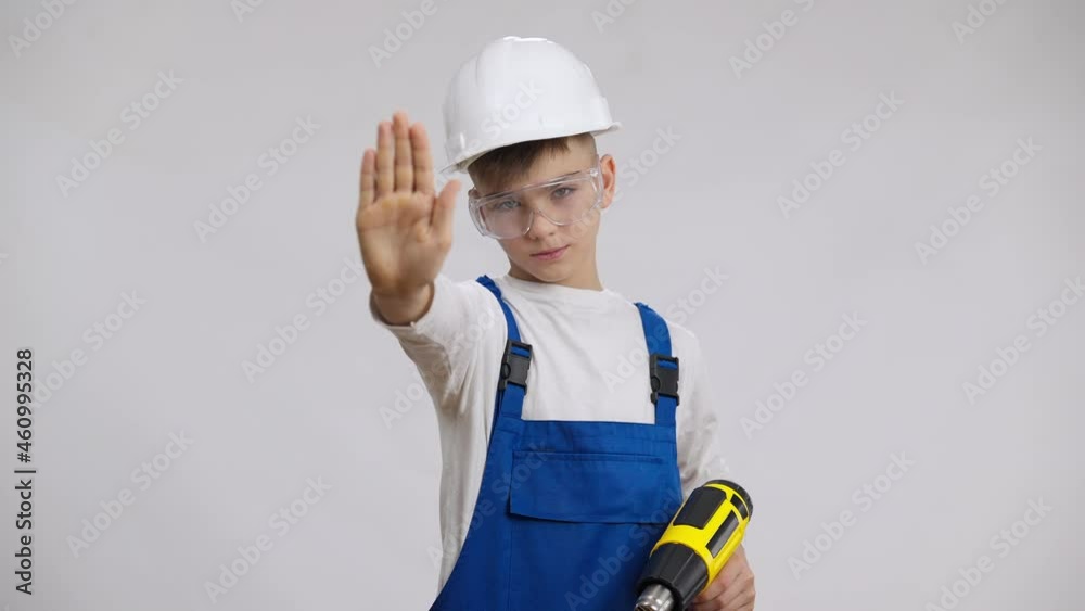Confident serious boy in hard hat and blue overalls stretching hand no gesture posing at white background with perforator. Portrait of cute Caucasian child choosing builder occupation. Work safety
