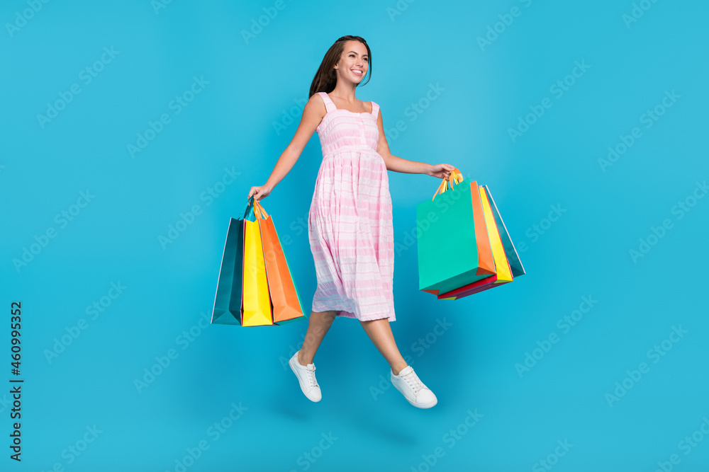 Full body photo of brunette haired stylish attractive beautiful woman jump hold bags go empty space sale isolated on blue color background Full body photo of brunette haired stylish attractive beautiful woman jump hold bags go empty space sale isolated on blue color background