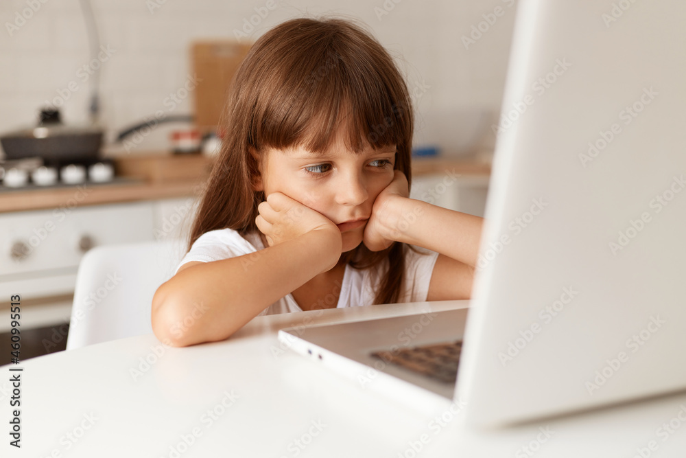 Indoor shot of cute bored dark haired female child sitting at table, looking at notebook screen with boring facial expression, having online lesson, keeping hands on face. Indoor shot of cute bored dark haired female child sitting at table, looking at notebook screen with boring facial expression, having online lesson, keeping hands on face.