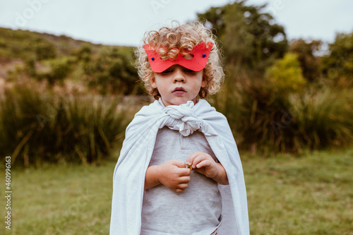 Cute blond boy with curly hair wearing mask and cape standing on meadow photo