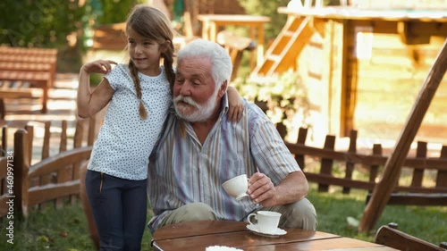 Sweet little girl hugs and kisses her grandfather in the yard while they spend time together. Elderly man drinks coffee in the yard and spends time with his granddaughter playing in the yard at home