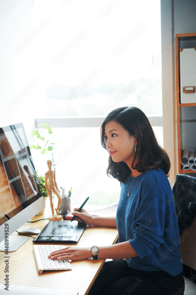 Content young Asian woman sitting at desk and working with graphics tablet while retouching photos for website Content young Asian woman sitting at desk and working with graphics tablet while retouching photos for website