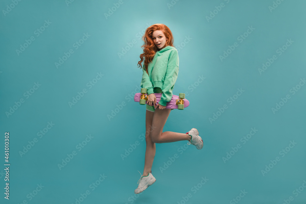 Active young girl with long curly hair in jump on blue background in studio. She is wearing a hoodie with shorts, holding skateboard. Leisure concept of youth, active lifestyle Active young girl with long curly hair in jump on blue background in studio. She is wearing a hoodie with shorts, holding skateboard. Leisure concept of youth, active lifestyle