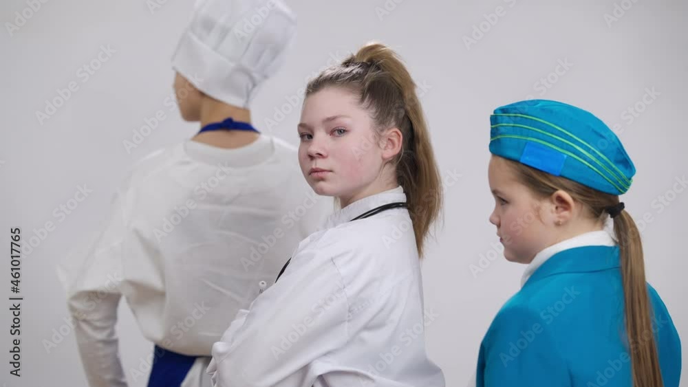 Confident serious Caucasian girl doctor turning crossing hands posing with blurred little stewardess and cook on both sides at white background. Portrait of child choosing medical profession Confident serious Caucasian girl doctor turning crossing hands posing with blurred little stewardess and cook on both sides at white background. Portrait of child choosing medical profession