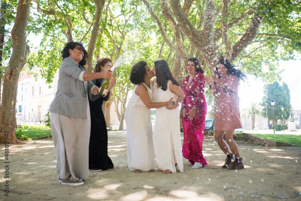Happy brides and guests celebrating wedding. Women of different nationalities in festive dresses laughing happily, applauding, opening champagne. Brides kissing. Wedding, LGBT, celebration concept
