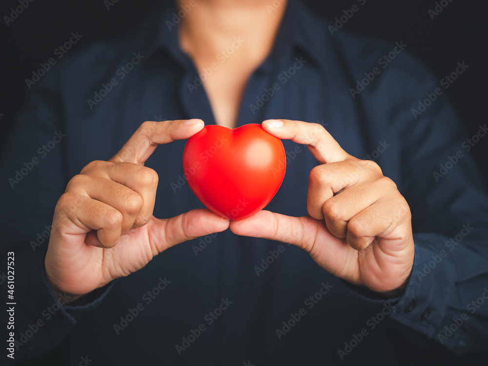 A young man in a blue shirt holding a red heart shape while standing with a black background in the studio. Midsection. Close-up photo. Space for text. Donate, love, and encouragement concept A young man in a blue shirt holding a red heart shape while standing with a black background in the studio. Midsection. Close-up photo. Space for text. Donate, love, and encouragement concept