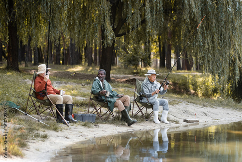 Multiethnic senior men with fishing rods holding thermo cups near lake