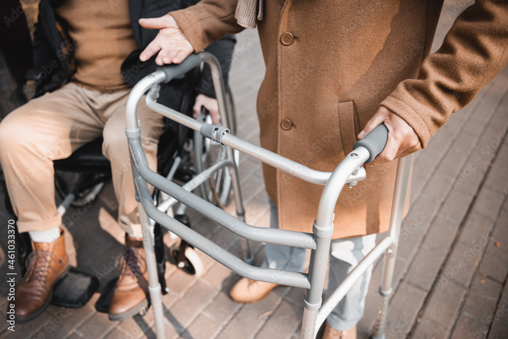 Obraz premium Cropped view of senior man with walking frame standing near friend in wheelchair outdoors