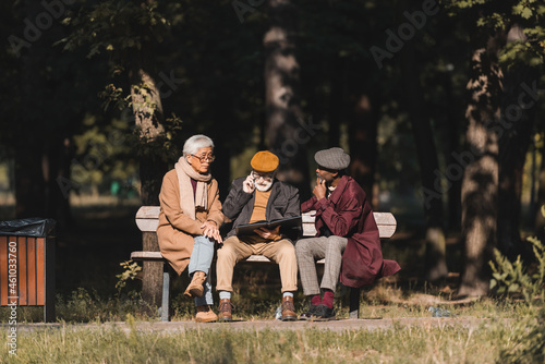 Senior interracial men sitting near friend with photo album talking on smartphone in park