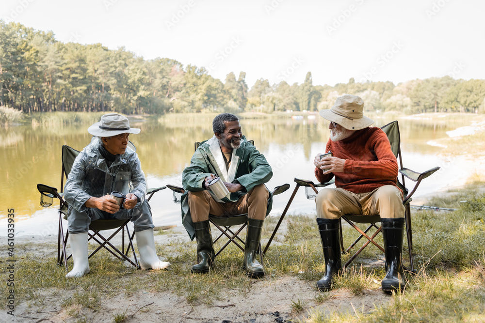 Cheerful multicultural men in fishing outfit holding thermo cups