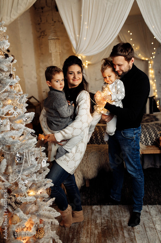 Joyful family with little kids standing near Christmas tree, celebrate winter holidays together at home. Beautiful parents with adorable son and daughter cuddling, looking at the camera, xmas concept
