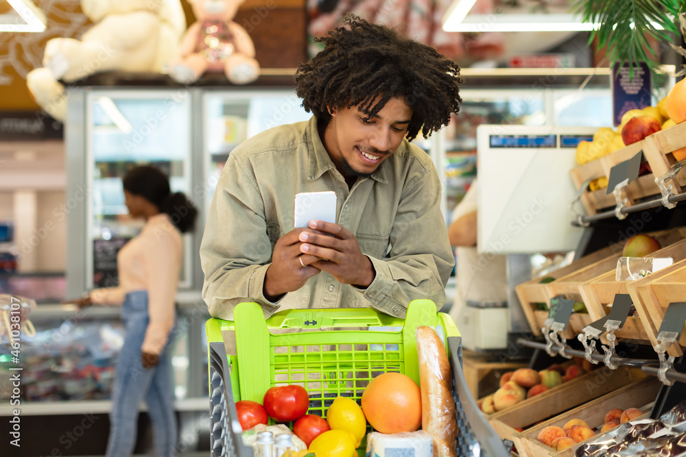 Black Man Using Smartphone While Shopping Groceries In Modern Supermarket