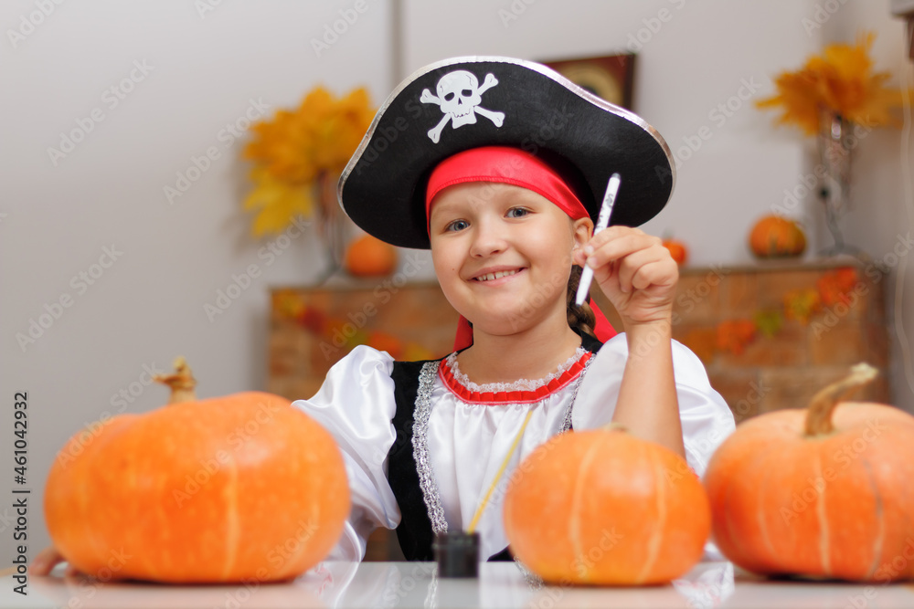 Celebrating Halloween at home. A little girl dressed as a pirate sits at the table and draws on a pumpkin. The child looks into the camera. - obrazy, fototapety, plakaty 