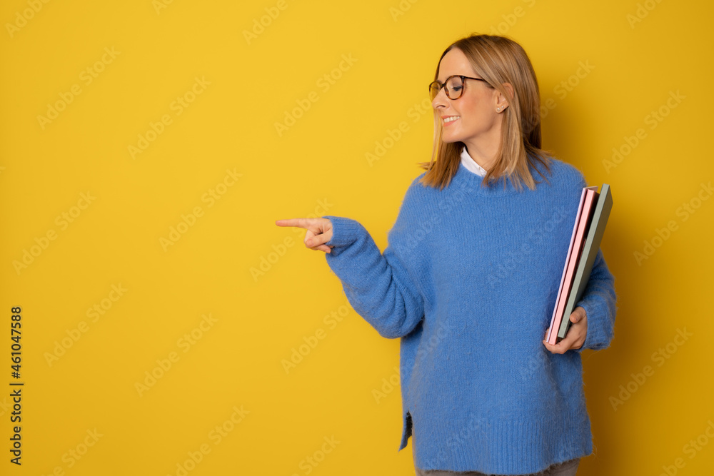 Portrait of happy young caucasian woman holding notebooks pointing finger side isolated on yellow background