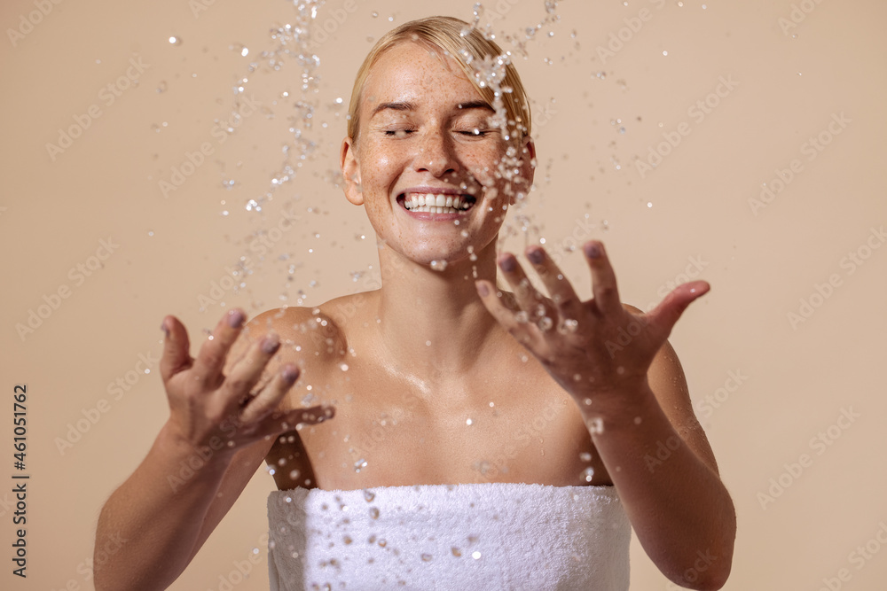 Happy woman wrapped in white towel splashing water on face in studio. Female with freckles on her skin enjoying skin care routine. Happy woman wrapped in white towel splashing water on face in studio. Female with freckles on her skin enjoying skin care routine.