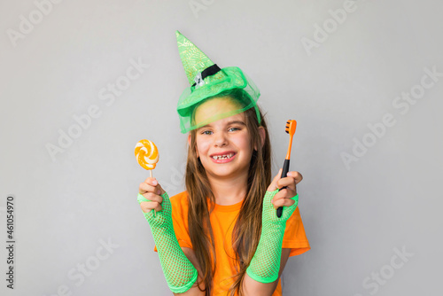 Happy Halloween. St. Patrick's Day.  Blonde girl kid in a witch costume with a candy and a toothbrush in her hands. Place for text, place for copying. Dental hygiene, children's health concept.