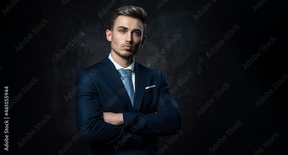 Conceptual portrait of a young, stylish business man, in a blue suit, hands on a cross, against a dark textured background.