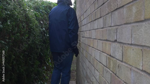 A man wearing denim jeans, a light jacket with hood up, and gloves, furtively walking next to an outside brick wall, then carefully peering around the corner. Burglar / intruder / private investigator photo