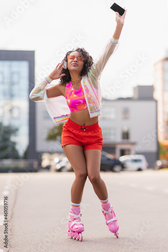 african american woman in wireless headphones and on roller skates singing while holding cellphone