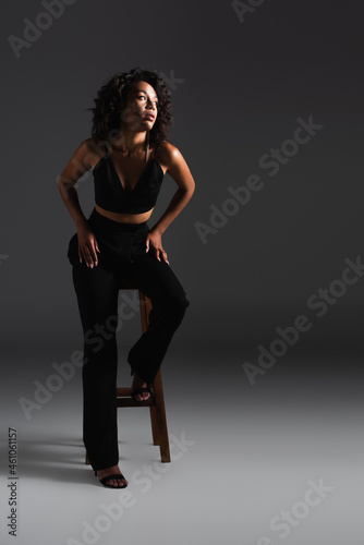 full length of curly african american woman in sexy outfit sitting on wooden stool on dark grey