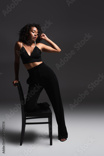 full length of brunette african american woman in trendy outfit leaning on chair on dark grey
