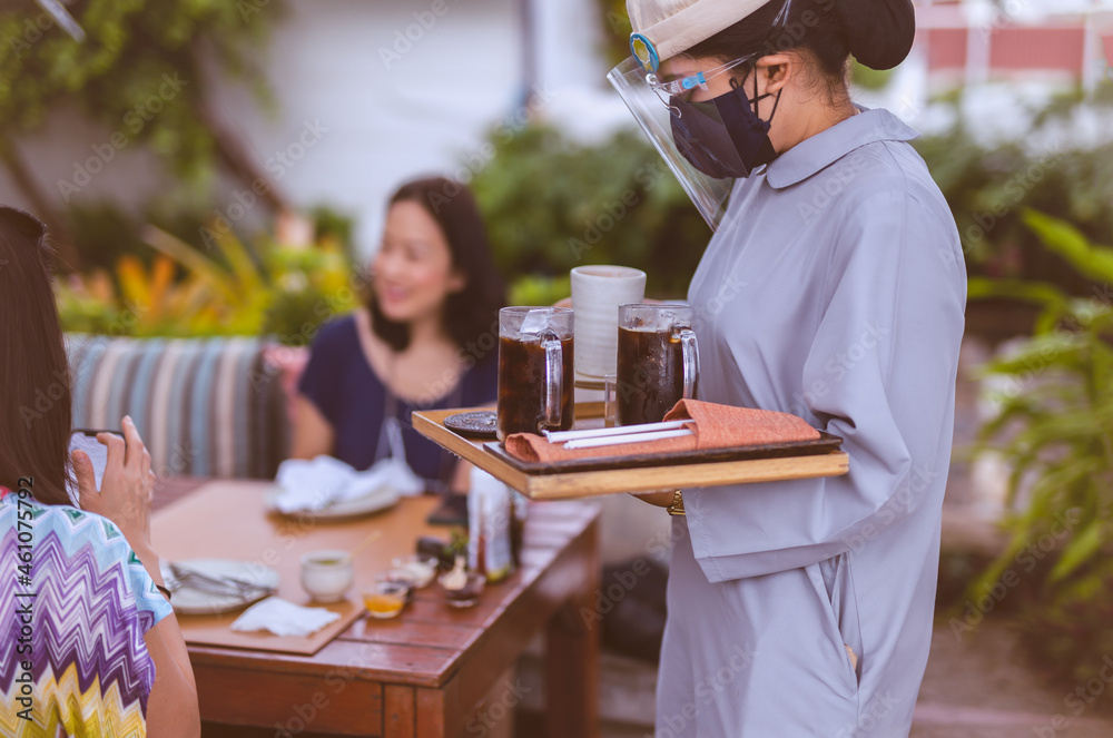 Female waiterss in face shield and mask serves iced coffee to customer. Female waiterss in face shield and mask serves iced coffee to customer.