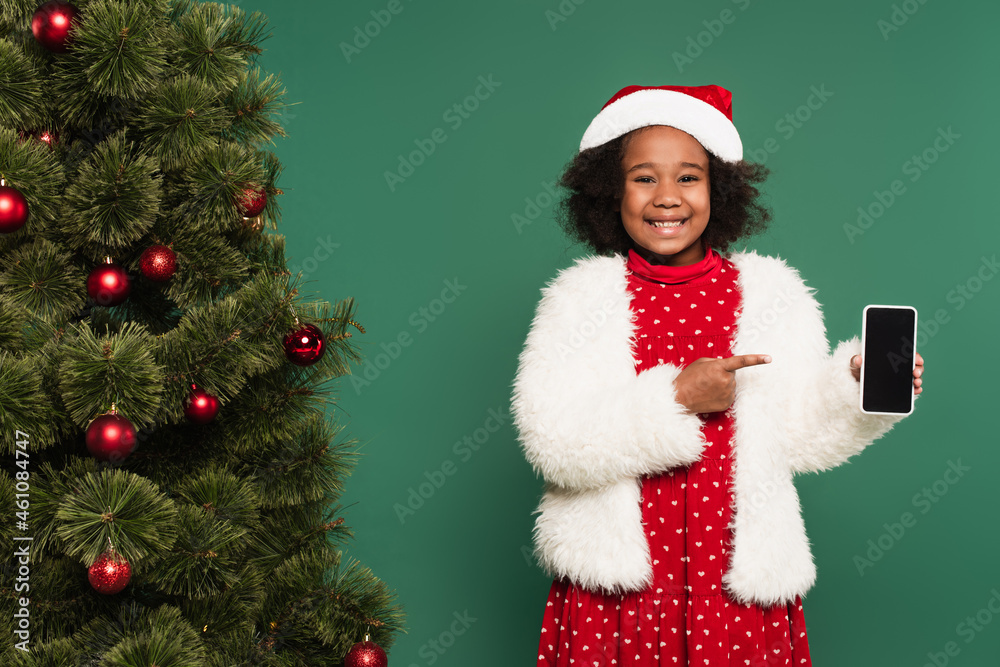 Positive african american girl in santa hat and fluffy jacket pointing at smartphone near decorated christmas tree isolated on green