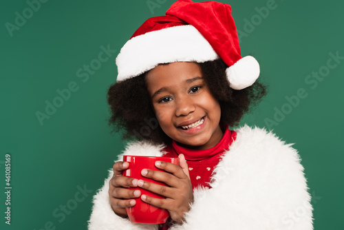 Positive african american kid in santa hat and fluffy jacket holding red cup isolated on green