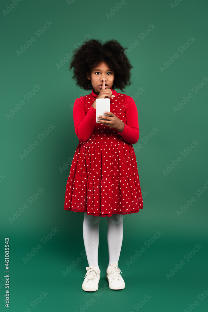 African american child in red dress showing secret gesture and using smartphone on green background African american child in red dress showing secret gesture and using smartphone on green background