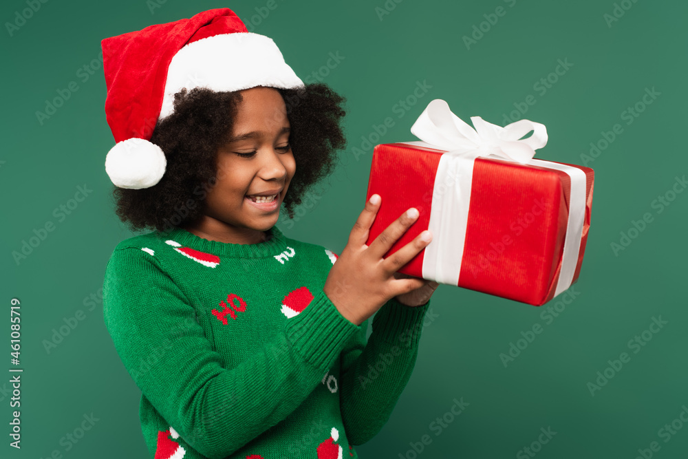 Positive african american girl in santa hat and sweater holding present isolated on green