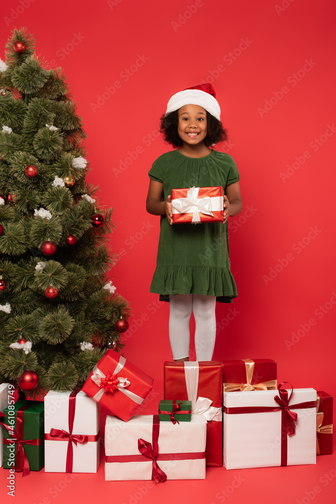 African american kid in santa hat holding present near decorated christmas tree on red background African american kid in santa hat holding present near decorated christmas tree on red background