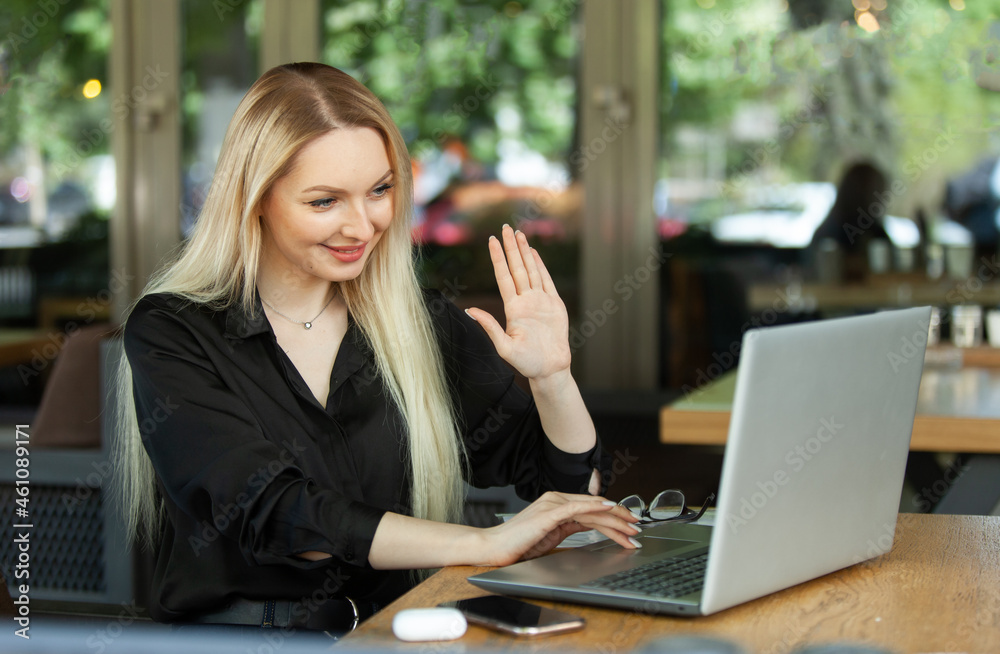Smiling business woman waving hand at laptop screen welcoming interlocutors by video conference while sitting at table in outdoor cafe Smiling business woman waving hand at laptop screen welcoming interlocutors by video conference while sitting at table in outdoor cafe