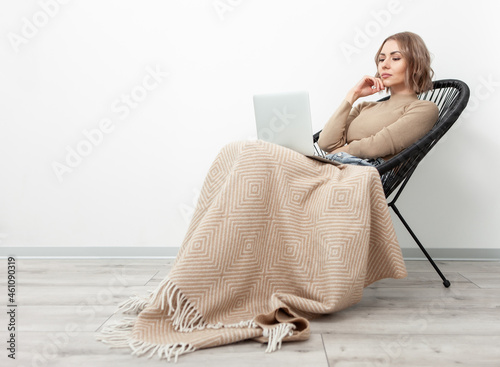 Beautiful woman with laptop sits on armchair, covered with blanket against the background of white wall. Leisure, lifestyle concept photo