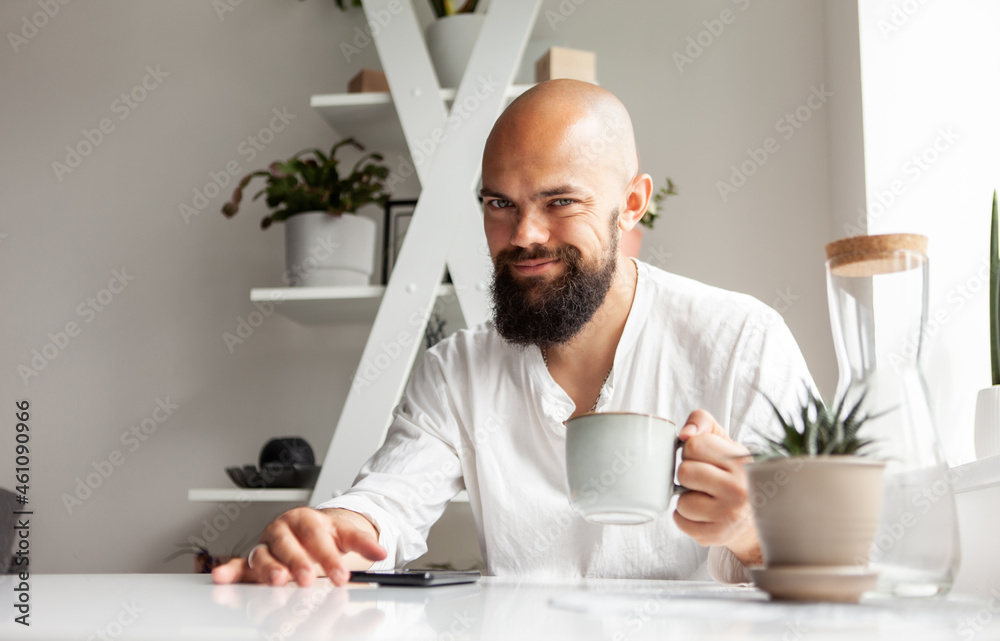 Handsome bald bearded man drinking coffee and using smartphone while sitting at the table in modern kitchen. Online work or leisure. Young male checks messages or reads news Handsome bald bearded man drinking coffee and using smartphone while sitting at the table in modern kitchen. Online work or leisure. Young male checks messages or reads news