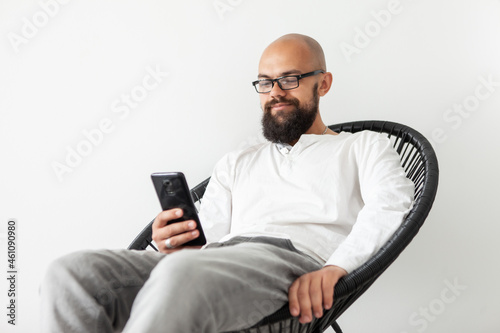 Handsome bald bearded man using smartphone while sitting in a chair in the living room. Online work or leisure. Young male checks messages or reads news