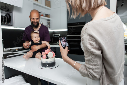 Fototapeta Naklejka Na Ścianę i Meble -  Birthday. Happy family concept. Young mother takes pictures of her daughter and husband with a cake in kitchen