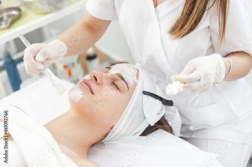 Cosmetological procedures. Beauty salon, doctor in gloves applies mask for beautiful woman with closed eyes with brush.