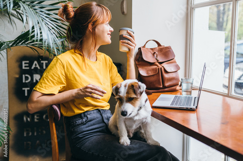 Young girl student sititng at table with computer and dog at work place. Pretty amazing female freelancer working at cafe with jack russell terrier. photo