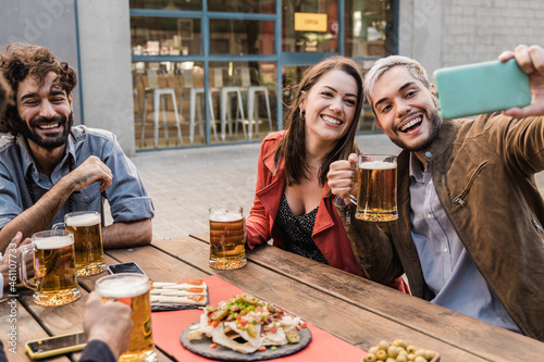 Young happy people having fun taking a selfie with mobile phone while drinking beer at brewery bar outdoor - Focus on girl face photo