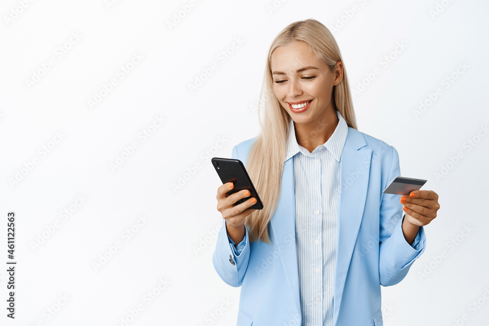 Smiling blond corporate woman enter info on mobile phone, holding credit card, wearing suit, standing over white background Smiling blond corporate woman enter info on mobile phone, holding credit card, wearing suit, standing over white background