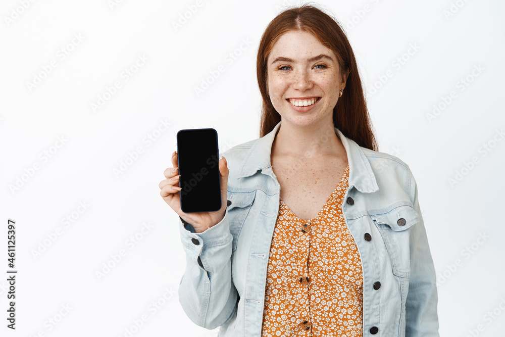 Beautiful young redhead woman showing her mobile phone screen, smiling pleased, recommending app, white background Beautiful young redhead woman showing her mobile phone screen, smiling pleased, recommending app, white background