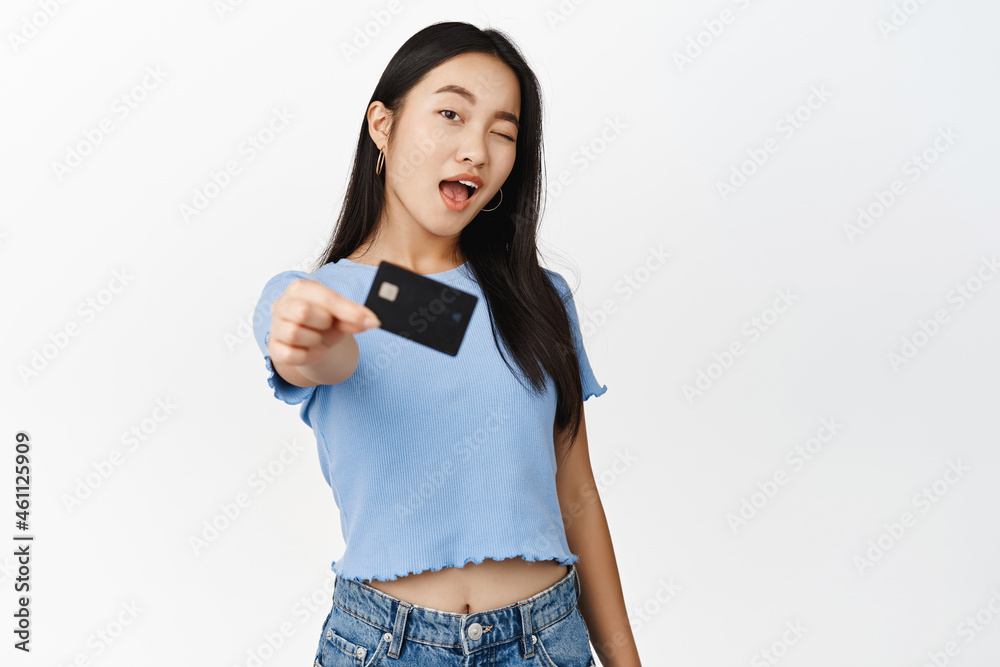 Happy modern asian girl showing credit card and winking at camera, recommending bank, standing over white background Happy modern asian girl showing credit card and winking at camera, recommending bank, standing over white background